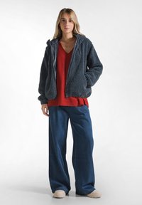 Giovane donna che indossa una giacca in pile grigio scuro, un maglione rosso a scollo a V, jeans a gamba larga blu e scarpe beige, in piedi davanti a uno sfondo bianco.