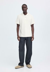 Polo blanc à manches courtes, patte de boutonnage à deux boutons, associé à un jean bleu foncé et des chaussures beige à enfiler sur un fond neutre.