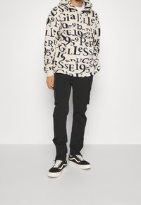 Kapuzenpullover in Creme mit blauen Textmustern, kombiniert mit schwarzen Cargohosen und schwarz-weißen Sneakers. Strukturierter Stoff, lässige Passform.