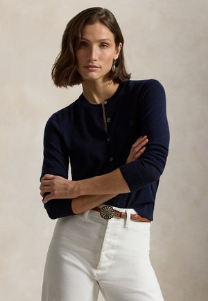 Femme aux cheveux courts et bruns portant un cardigan bleu marine boutonné, un pantalon blanc, une ceinture en cuir marron avec une boucle décorative, debout, les bras croisés.