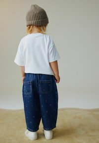 Camiseta blanca de manga corta, pantalones vaqueros azules con lunares y zapatillas blancas. Se lleva un gorro beige de punto en la cabeza.