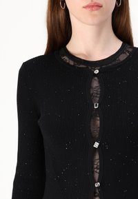 Cardigan nero a coste con pannello in pizzo sul collo, decorato con bottoni in cristallo geometrici e un sottile luccichio in tutto il capo.