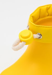 Botte en caoutchouc imperméable jaune avec un bout rond, un col matelassé et un accent en corde blanche fixé par un bouton jaune et des œillets en métal.