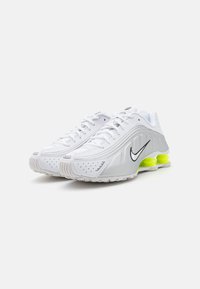 Baltos sportinės bateliai su lygia sintetine viršutine dalimi, subtiliomis pilkomis detalėmis, juodu Nike logotipu ir ryškiai geltonu padu kulno srityje.