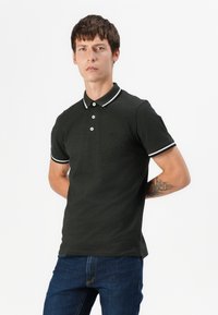 Polo shirt vert foncé avec un col, des bordures blanches et trois boutons. Manches courtes et design ajusté sans logos visibles. Porté avec un jean bleu.