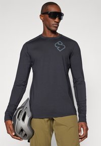 Zwart sportshirt met lange mouwen en een subtiel logo, gecombineerd met olijfgroene broek en een grijze helm met ventilatiegaten.