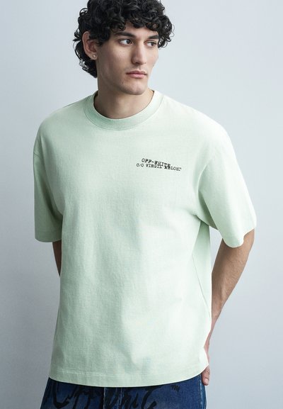 Jeune homme aux cheveux bouclés portant un t-shirt Off-White vert pâle et un pantalon bleu à motifs, regardant vers sa droite sur un fond clair.