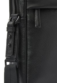 Zwarte leren tas met een gestructureerde oppervlakte, voorzien van een versterkte riembevestiging en mat afgewerkte hardware. Ritsafsluiting langs de rand.