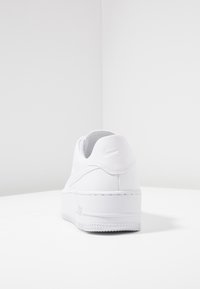 Białe sneakersy Nike Air Force 1 pokazane z tyłu, z wytłoczonymi detalami logo i grubą gumową podeszwą na białej powierzchni.