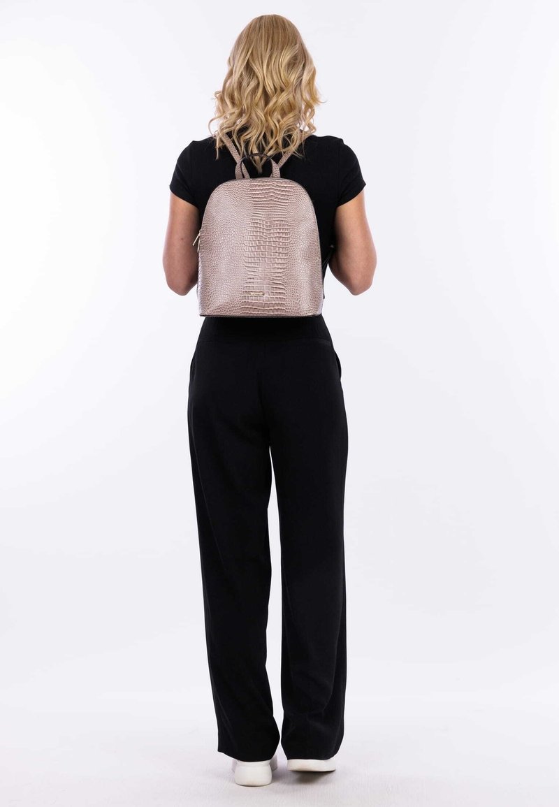 Femme aux cheveux blonds portant une tenue noire et des chaussures blanches, avec un sac à dos rose texturé, debout sur un fond blanc.