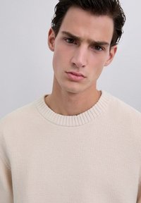 Lichtbeige gebreide trui met een ribgebreide ronde hals. De textuur is iets chunky, met een zachte, geweven patroon. Close-up van het kledingstuk.