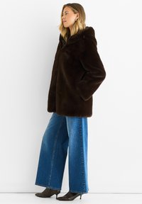 Brown faux fur coat with a hood, loose fit, side pockets; paired with wide-leg blue jeans and pointed black textured ankle boots.