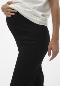 Svarta stretchbyxor med en slät textur, designade för komfort, tillsammans med en vit åtsittande skjorta. Synlig magkurva framhävd.