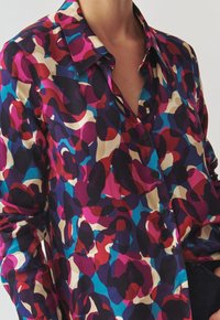 Camicia colorata con bottone, caratterizzata da un motivo floreale in tonalità di viola, rosa, blu e beige; completata da un colletto classico e maniche lunghe.