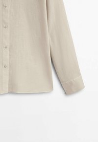 Chemise beige clair à manches longues avec boutons sur le devant, montrée de la moitié du torse jusqu'au poignet sur un fond blanc.
