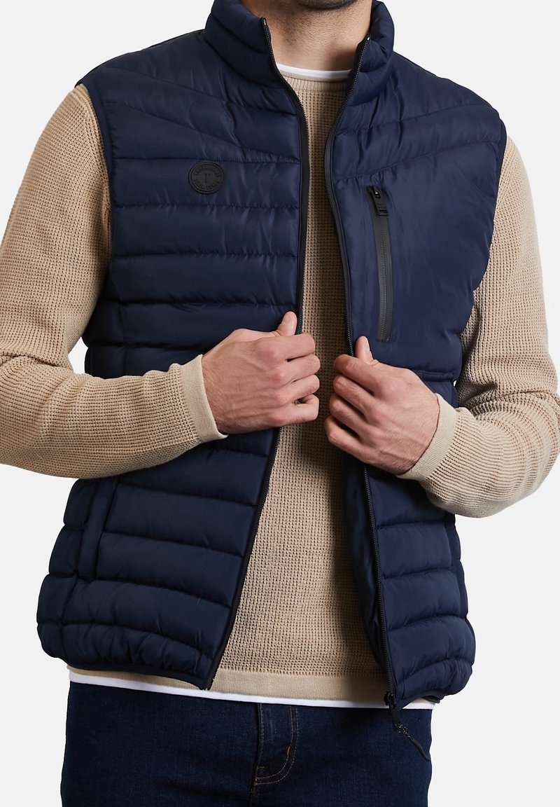 Navyblauer Steppwesten mit hohem Kragen, horizontalem Steppmuster und einer Reißverschlusstasche. Über einem beigen Strickpullover getragen.