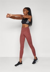 Soutien-gorge de sport noir avec des rayures blanches ; leggings rouges texturés ; chaussures de sport noires. La personne s'étire avec les bras tendus.