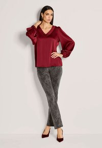 Bordeauxfarbige Satinbluse mit V-Ausschnitt und Puffärmeln, kombiniert mit grauen gemusterten Hosen. Das Model steht in roten Absätzen vor einem neutralen Hintergrund.