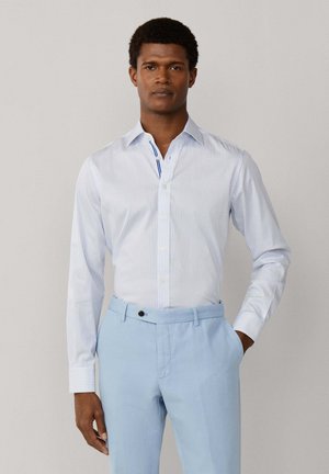 Uomo che indossa una camicia a righe azzurro chiaro a maniche lunghe infilata in un pantalone elegante azzurro chiaro, in piedi con una mano in tasca.