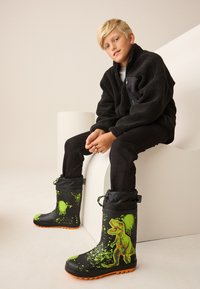 Stivali impermeabili neri con design di T-Rex in macchie verdi, suola arancione e parte superiore trapuntata, con chiusura a coulisse e superficie testurizzata.