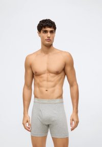 Muž bez trička s krátkými tmavými vlasy, oblečený v světle šedých boxerkách Calvin Klein, stojící před jednoduchým bílým pozadím.