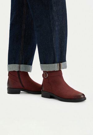 Knöchelhohe dunkelrote Lederstiefel mit seitlichen Reißverschlüssen und Schnallen, getragen zu hochgekrempelten dunkelblauen Jeans.