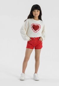 Bela majica s kapuco z rdečo bleščečo srčno zasnovo, v kombinaciji z rdečimi jeans kratkimi hlačami z raztrganimi robovi. Model nosi bele superge.