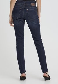 Mujer vistiendo jeans ajustados azul oscuro y sandalias negras de tacón alto, vista desde atrás contra un fondo liso.