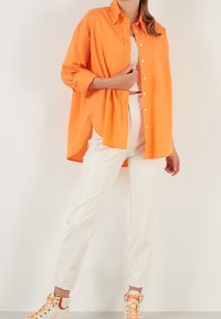 Person klädd i en klar orange oversized skjorta med knappar, vita något korta byxor och vita sneakers med orange snören mot en vit bakgrund.