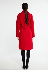 Cappotto rosso in lana con cintura, maniche lunghe e spacco posteriore. Texture liscia e design fino al ginocchio. Indossato con leggings neri e stivali con tacco.