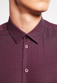 Chemise bordeaux à boutons, présentant un motif géométrique, un col pointu et des boutons violets. Texture lisse avec une coupe ajustée.