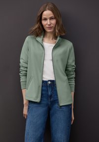 Giacca verde chiaro con zip, collo alto, maniche lunghe e tasche laterali, abbinata a jeans blu, su uno sfondo scuro.