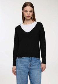 Maglione nero a scollo a V con maniche lunghe indossato sopra una camicia bianca. Il design presenta una vestibilità ampia e polsini a coste, abbinati a jeans azzurri chiari.