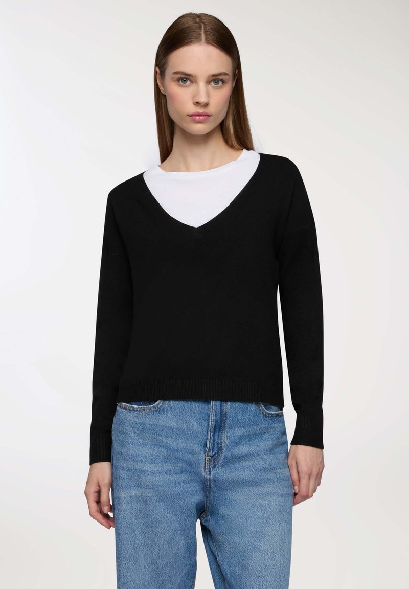 Maglione nero a scollo a V con maniche lunghe indossato sopra una camicia bianca. Il design presenta una vestibilità ampia e polsini a coste, abbinati a jeans azzurri chiari.