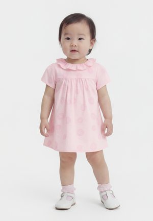 Väikelaps, kes kannab roosat kleiti volditud kraega, roosasid sokke ja valgeid kingi, seisab valgel taustal.