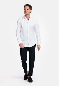 Chemise blanche à manches longues, associée à un pantalon slim-fit bleu marine et des chaussures noires. Tissu lisse, sans motifs visibles.