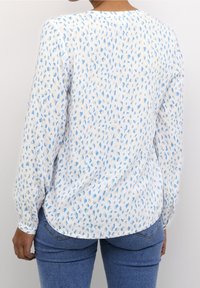 Chemise blanche à manches longues avec un motif dispersé de motifs bleus, col rond et ourlet légèrement arrondi, associée à un jean en denim bleu.
