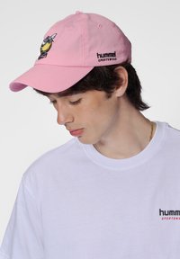 En rosa bomull baseballkeps har en broderad grafisk design och texten "hummel sportswear" i svart, bärs med en vit t-shirt.