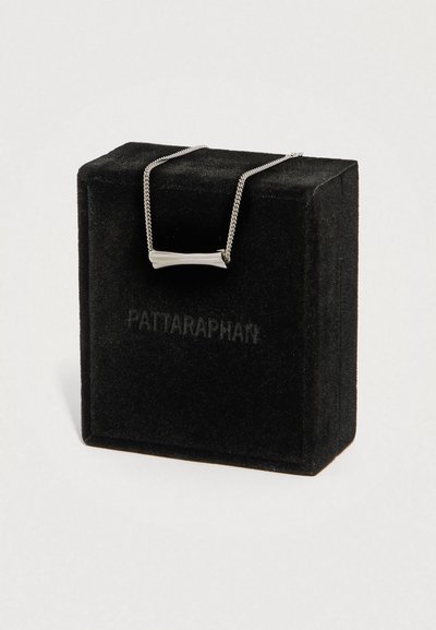 Ristkülikukujuline hõbedane ripats kettal, mis on esitatud musta sametkarbi kohal, kus on õrnalt reljeefne tekst.