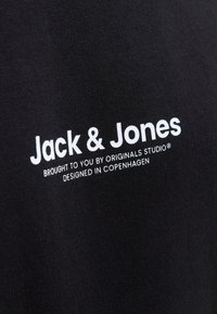 Černé bavlněné tričko s bílým nápisem: "Jack & Jones, přineseno Originals Studio, navrženo v Kodani." Hladká textura.