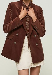Hnědý kordový dvouřadý blejzr se stříbrnými knoflíky, s klopami a předními kapsami, vrstvený přes košili s květinovým vzorem.
