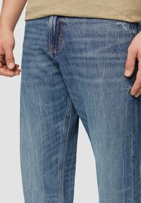 Jean en denim bleu clair avec une texture décolorée, présentant des coutures contrastantes orange, un design à cinq poches et une coupe décontractée.