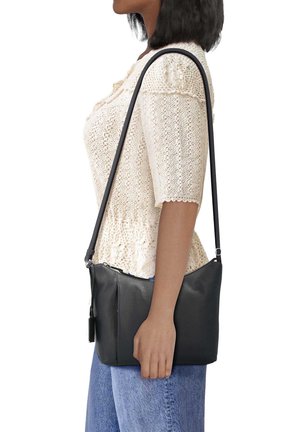 Femme portant un haut tricoté crème et un jean bleu, tenant un sac bandoulière en cuir noir avec une longue sangle sur l'épaule.