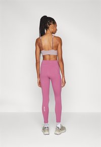 Hellblauer Sport-BH mit verstellbaren Trägern, kombiniert mit hochgeschnittenen pinken Leggings, die ein weißes Logo zeigen. Model mit grauen Turnschuhen.