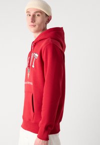 Sudadera de algodón roja con un bolsillo frontal, puños acanalados y letras con marca en el pecho. Combinada con pantalones color crema y un gorro beige.