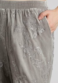 Acercamiento de una mano en el bolsillo de pantalones de terciopelo gris claro con bordado de lentejuelas florales y cintura elástica.