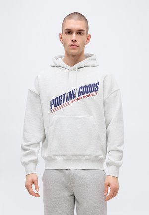 Jeune homme portant un sweat à capuche gris clair avec le texte "SPORTING GOODS BJÖRN BORG" et un pantalon de survêtement assorti gris clair, debout devant un fond uni.
