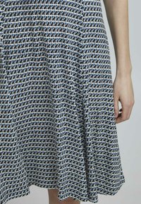 Tryckt klänning med geometriskt mönster i marinblått och vitt, med en utsvängd silhuett och mjuk textur. Synlig hand nära fållen.