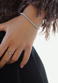 Mano con un anillo de plata que muestra una pequeña piedra clara en el dedo anular y una pulsera con cuentas blancas espaciadas de manera uniforme en la muñeca.