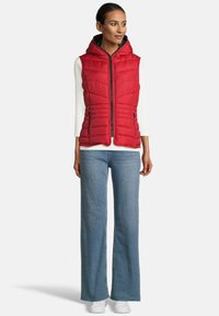 Vrouw in een rood mouwloos gewatteerd vest met capuchon, over een witte lange mouw top, met blauwe jeans en witte sneakers, staand in een vooraanzicht.